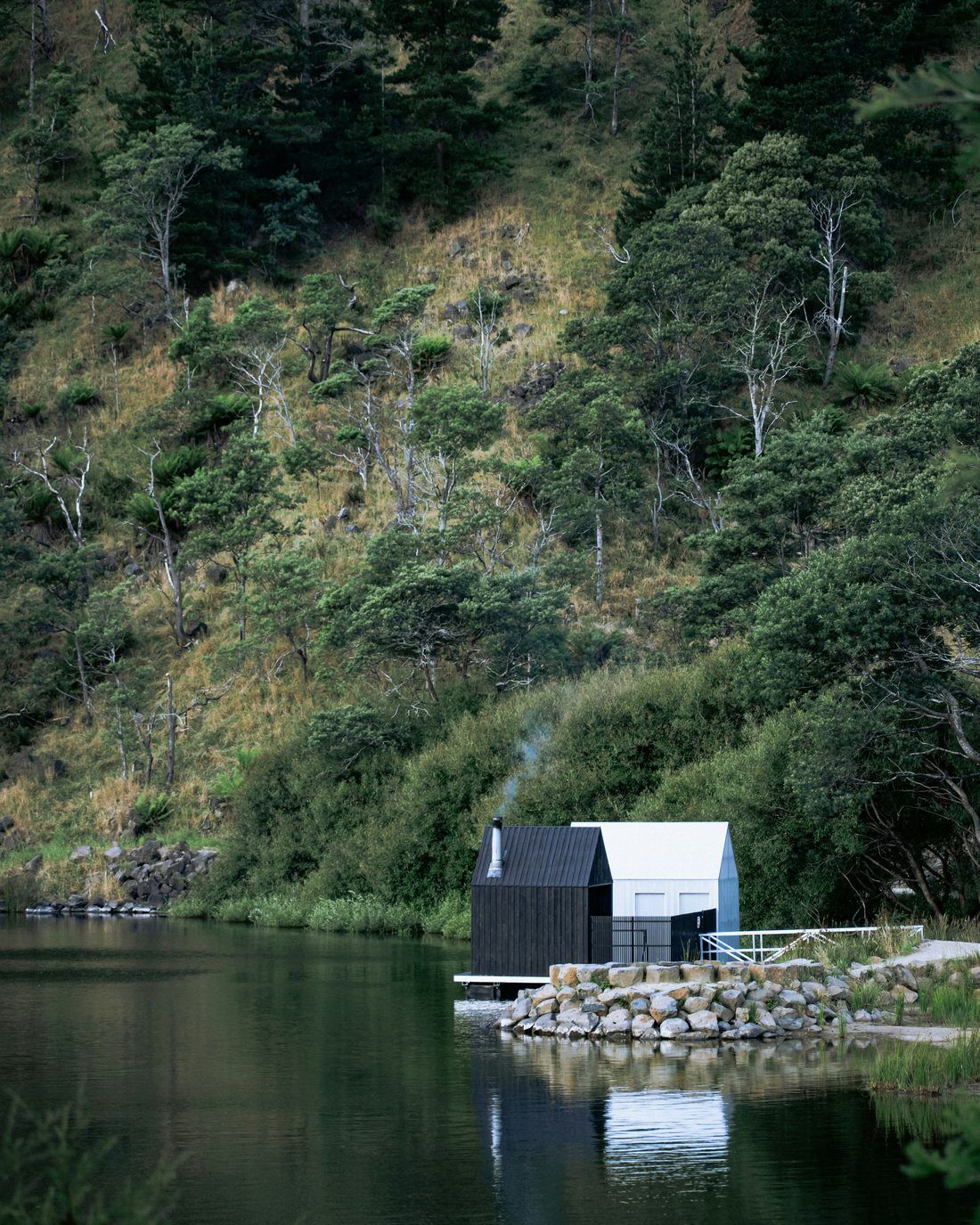 A small black timber sauna and a white hut at the edge of a still lake, smoke rising, below a steep green hillside.