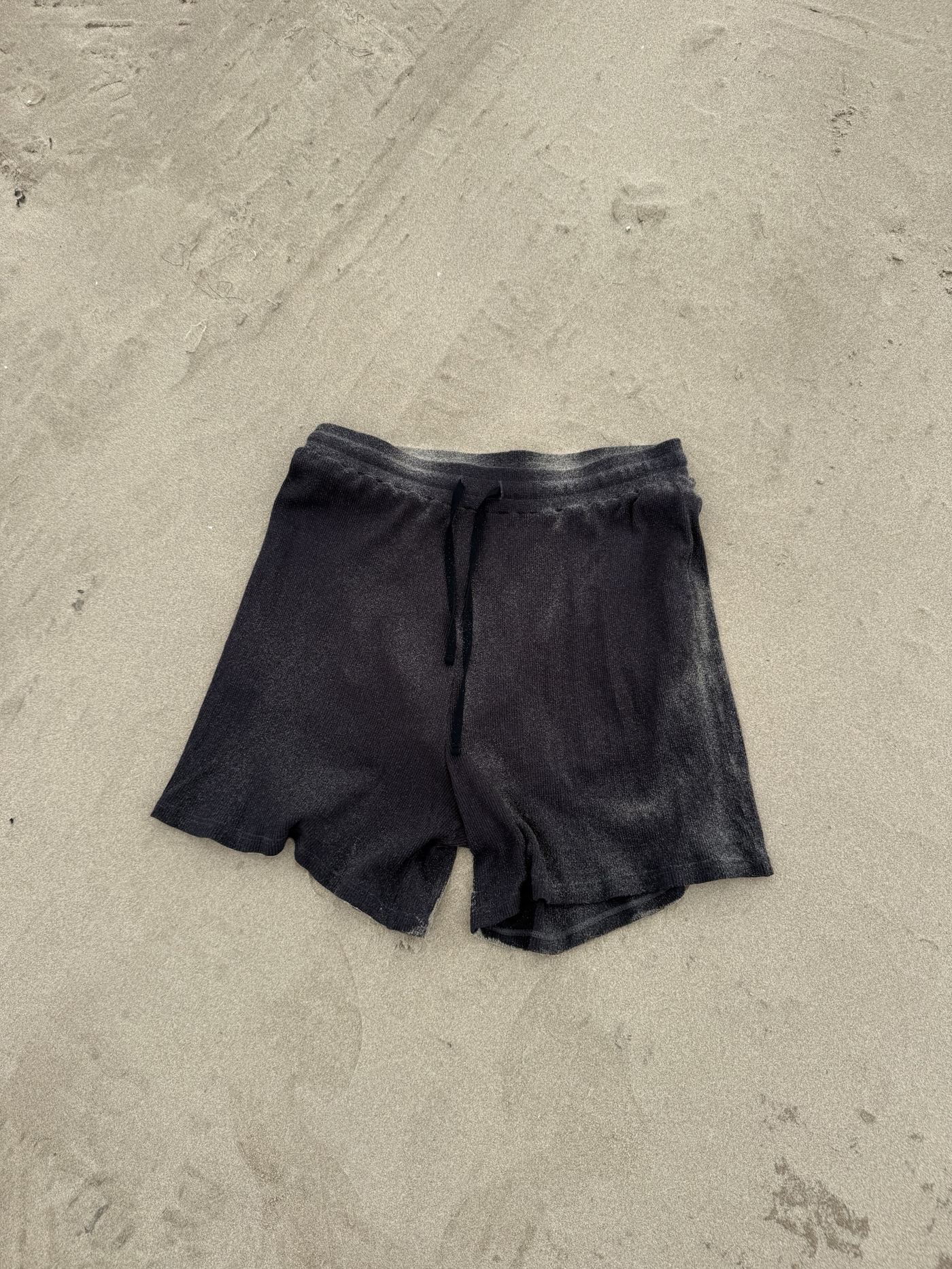 A charcoal waffle knit short with a drawstring, laid flat on sand.